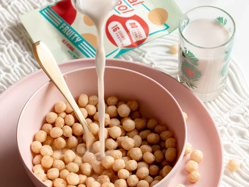 Milk being poured into a pink bowl of Schoolyard Snacks cereal with a spoon and cereal pack nearby.
